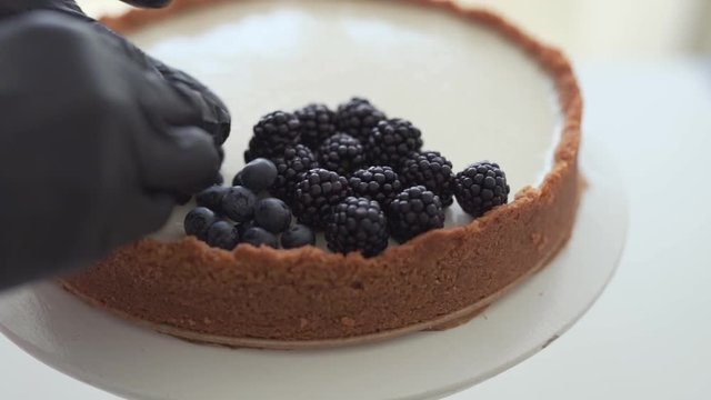 Close-up scene of a cook in a black gloves who has just made a cheesecake and now has decorating it with berries : blackberries and bilberries. 