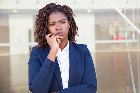 Focused pensive leader thinking outside. Serious young black business woman standing at outdoor glass wall, touching chin and looking away into distance. Thinking concept