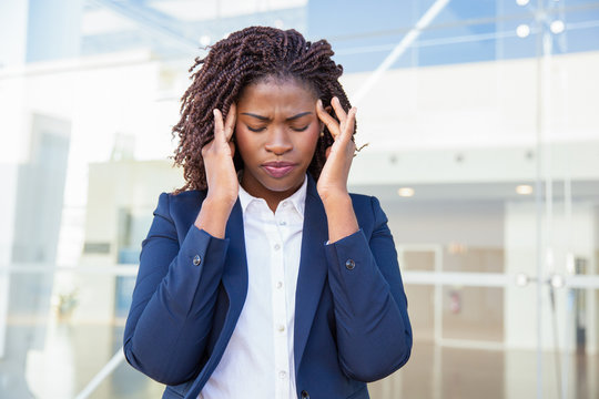 Stressed tired female office employee suffering from headache. Young black business woman with closed eyes and pain face standing outside, touching head and temples. Fatigue or sickness concept