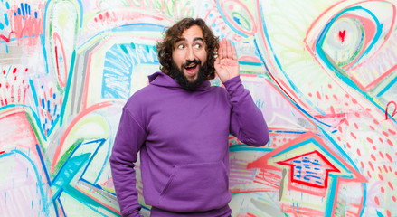 young bearded crazy man smiling, looking curiously to the side, trying to listen to gossip or overhearing a secret against graffiti wall