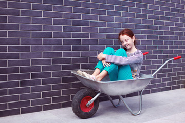 Teen girl sitting in a construction trolley on a brick wall background and smiling. Close-up. Construction concept. Copy space.