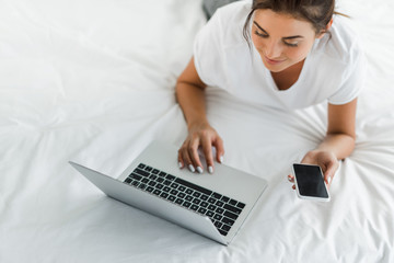attractive girl using smartphone and laptop in bed in the morning