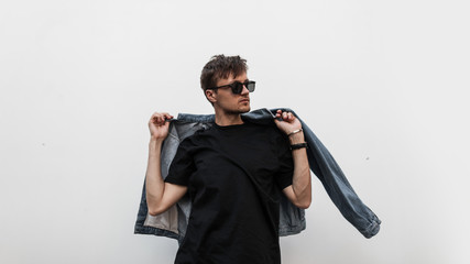 Cool modern young hipster man with a stylish hairstyle in sunglasses in a black T-shirt puts on a...