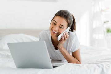 attractive smiling girl talking on smartphone and using laptop in bed in the morning