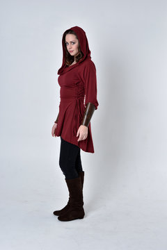 full length portrait of a brunette girl wearing a red fantasy tunic with hood. Standing pose on a white studio background