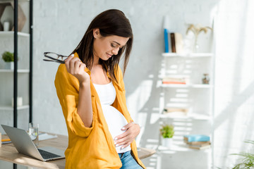 happy pregnant freelancer touching belly while sitting in home office