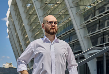 A horizontal shot of a bald young man in glasses with a beard in a white shirt is standing against the background of building in city. Busy young man. Arms on sides, bent at the elbows. Looking.