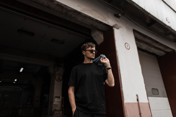Cool nice young man in stylish sunglasses in a vintage blue denim jacket in a black T-shirt poses...