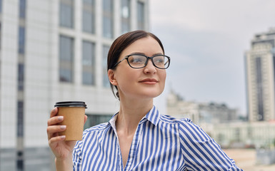 A nice looking busy girl stands against the backdrop of the city, holds a brown cup coffe in her hand and looks away with smile. City view, business.