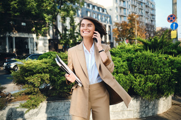 Young cheerful businesswoman in beige suit with laptop happily talking on cellphone on street