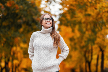Model of a happy European young woman in a knitted fashionable white sweater in stylish glasses with brown hair with a beautiful smile relaxes in the park on an autumn day. Positive hipster girl.