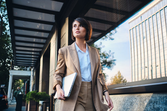 Young attractive stylish businesswoman with laptop thoughtfully walking on city street