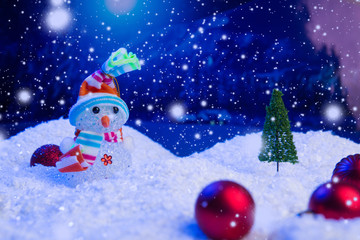 Snow Man with Christmas balls on snow over fir-tree, night sky and moon. Shallow depth of field. Christmas background. Fairy tale. Macro. Artificial magic dreamy world.
