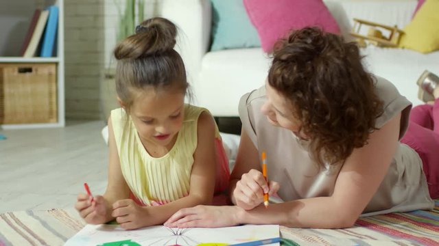Happy mother and cute little daughter drawing together on paper and discussing picture while lying on the floor at home