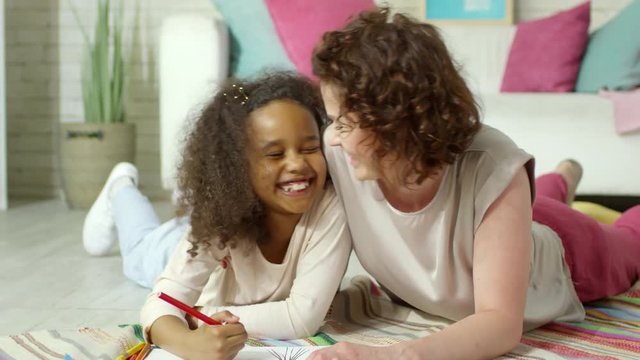Caucasian female teacher smiling and hugging cute African girl while lying on the floor in kindergarten and drawing picture together