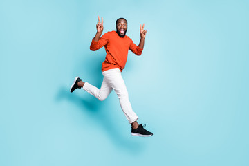 Full length body size photo of cheerful positive casual man running jumping mixed-race wearing white pants trousers sneakers showing double v-sign isolated vivid blue color background