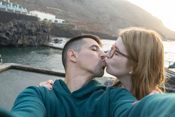 Lovers kissing with passion taking selfie in natural pool. Caucasian blonde girlfriend with glasses kiss latin boyfriend at sunset. Adolescent Couple Having Romantic moment in the coast.