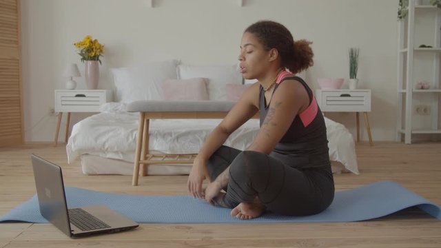 Positive sporty african american woman consulting with fitness trainer online via laptop while sitting on exercise mat at home. Cheerful female using online personal fitness trainer service on laptop.