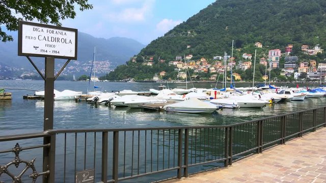 road sign of the pedestrian bridge that crosses Lake Como with the written words 'Diga Foranea, Piero Caldirola, physicist 1914-1984' with motor boats in the background