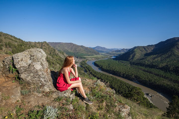 Naklejka na ściany i meble Portrait of a woman in a red dress on top of a mountain on the background of the river flowing between the rocks.