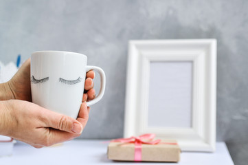 Beauty eyelash concept: women's hands hold a white Cup with eyelash extension decor on the table with other stylish things and tools on a gray background