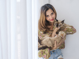 Smiling Asian woman holding her cat with window light at home.