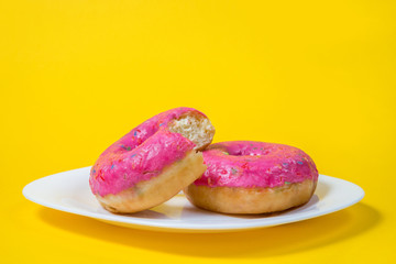 Two sweet half-eaten pink doughnuts on a white plate isolated on a yellow background. Minimal summer concept. Flat lay. The concept of unhealthy eating and weight gain. Harmful fast food. Copy space