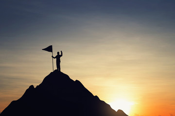 Silhouette of businessman holding a flag on top mountain, sky and sun light background. Business success and goal concept. - obrazy, fototapety, plakaty