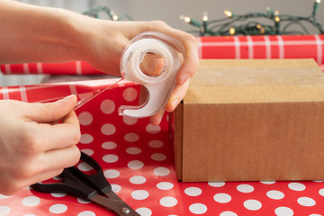 Female person pulling clear thin sticky tape from a dispenser and using it to stick polka dot wrapping paper onto a plain brown cardboard box
