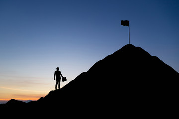 Silhouette of businessman looking at a flag on top mountain, sky and sun light background. Business and goal concept.