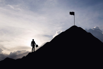 Silhouette of businessman looking at a flag on top mountain, sky and sun light background. Business and goal concept.