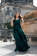 Fototapeta premium Beautiful girl with a long curly hair and make up in a long evening dress in motion on a walk in european streets. Sunny, summer weather .