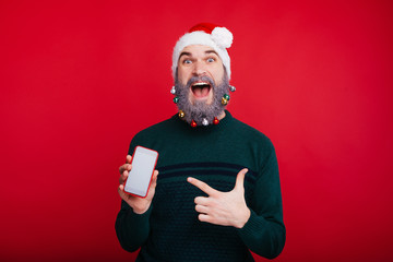 Amazed man with decorated beard and wearing santa claus hat pointing at smartphone screen