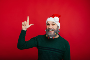 Cheerful man with decorated beard pointing up at copyspace over red background