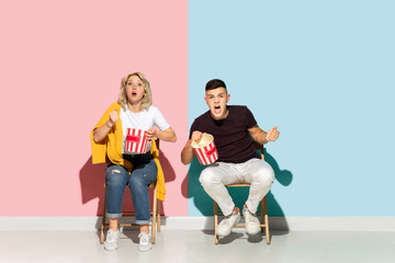 Fototapeta na wymiar Young emotional man and woman in bright casual clothes posing on pink and blue background. Concept of human emotions, facial expession, relations, ad. Beautiful couple watching cinema with popcorn.