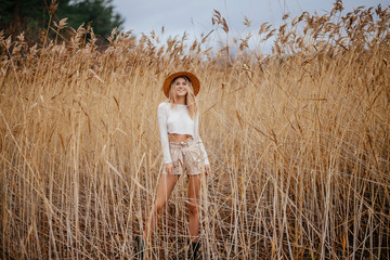 Excited blonde happy safari style girl in a straw hat walks on nature among the reeds. Lifestyle portrait of a young woman standing in yellow reed field in sunny autumn day countryside. 