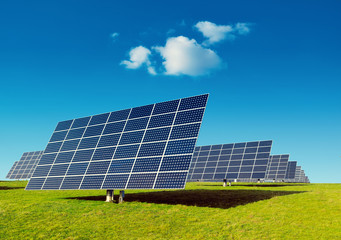 Rows of solar panels on a green grass field under blue sky with a small cloud