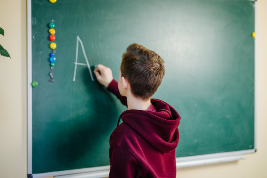 Student of higer school is standing in dark hoodie near the green blackboard in the classroom. Teenage Boy is writing alphabet on the board. School education concept. - Powered by Adobe