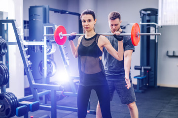 Sport, bodybuilding, lifestyle and people concept - young man and woman with barbell flexing muscles and making shoulder press squat in gym. Trainer and fitness girl are having workout at gym.