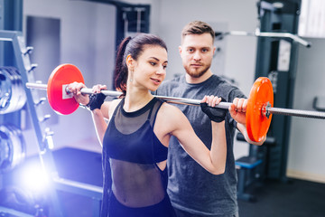 Sport, bodybuilding, lifestyle and people concept - young man and woman with barbell flexing muscles and making shoulder press squat in gym. Trainer and fitness girl are having workout at gym.