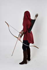 full length portrait of a brunette girl wearing a red fantasy tunic with hood, holding a bow and...