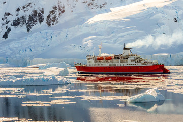 Red cruise steamboat drifting among the icebergs with huge rock and glacier in background, Neco bay, Antarctica - obrazy, fototapety, plakaty