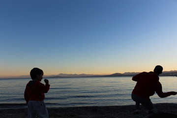 Father and son throwing stones to the sea from the shore after sunset one summer afternoon