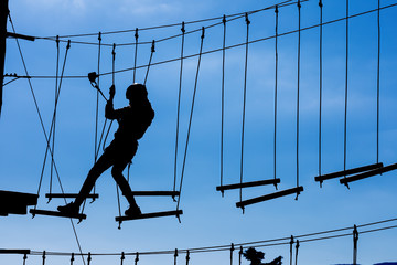 Silhouette of young girl in helmet climbing on high rope course against blue sky. Wooden bars tied to ropes, part of obstacle course in adventure park