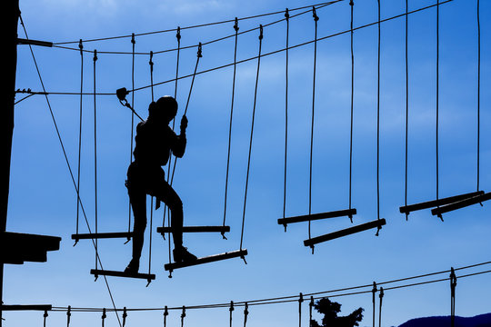 Silhouette of young girl in helmet climbing on high rope course against blue sky. Wooden bars tied to ropes, part of obstacle course in adventure park