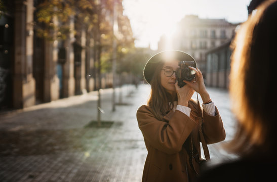 Outdoor smiling lifestyle portrait of pretty young woman having fun in sun city Europe autumn with camera travel photo of photographer Making pictures in hipster glasses and hat with girlfriends