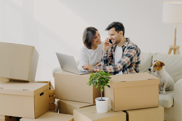 Happy husband and wife move in new bought house, buy furniture online, use modern technology, man surfs internet via laptop and talk over phone, sits on sofa with girlfriend and dog, many carton boxes - obrazy, fototapety, plakaty