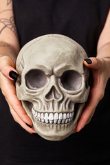 Tattooed hands of a woman in a black watch and clothes are holding a realistic model of a human skull with teeth. Close-up shot.