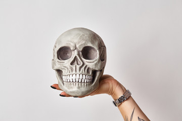 Tattooed hand of a woman in a black watch is holding a realistic model of a human skull with teeth isolated on white. Close-up shot.