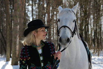 Obraz premium Beautiful young blond woman in colorful jacket, hat and black trousers looks with love on white horse in sunny winter day in the forest 